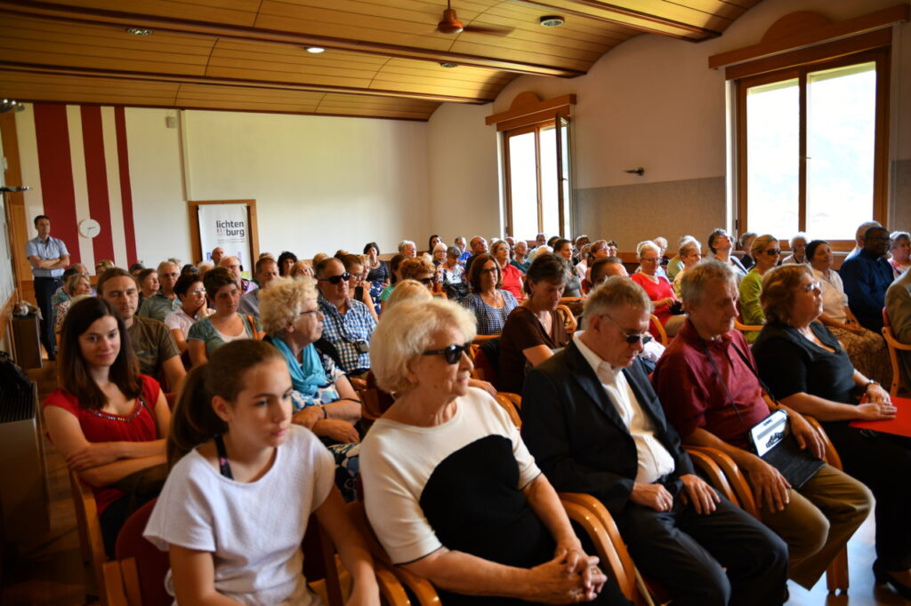Eine große Gruppe von Menschen sitzt in einem Veranstaltungsraum und lauscht aufmerksam.