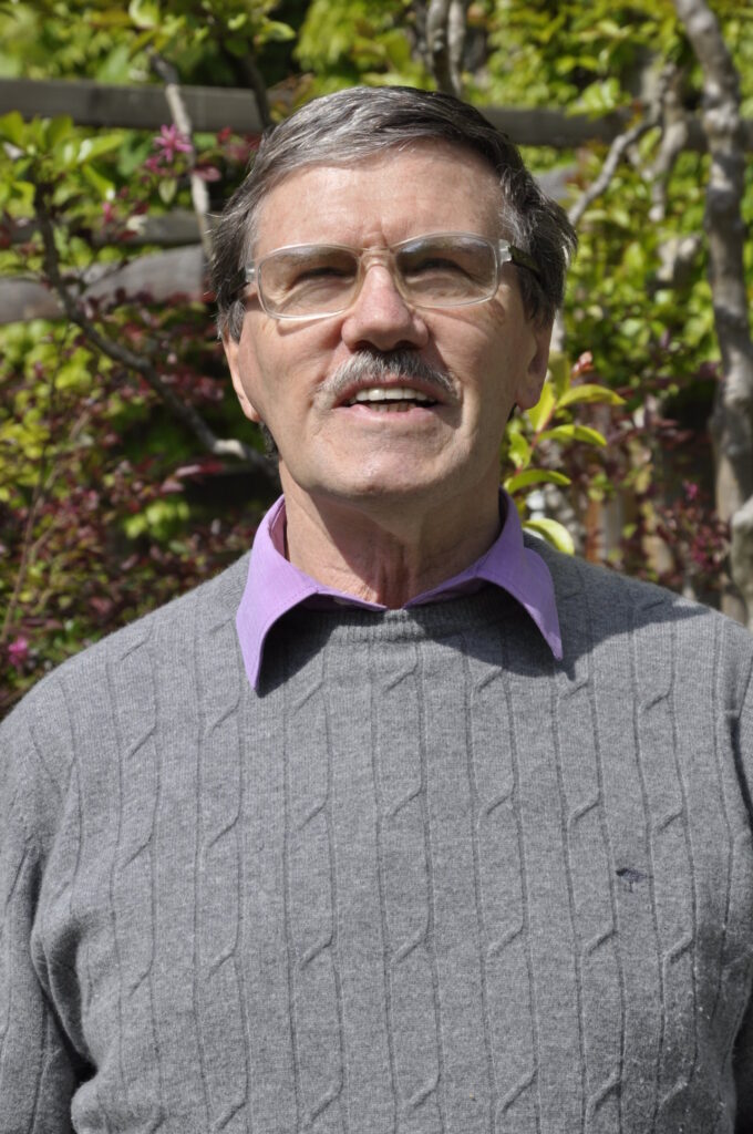 Ein älterer Mann mit Brille und grauem Pullover steht lächelnd im Freien vor grünen Büschen.