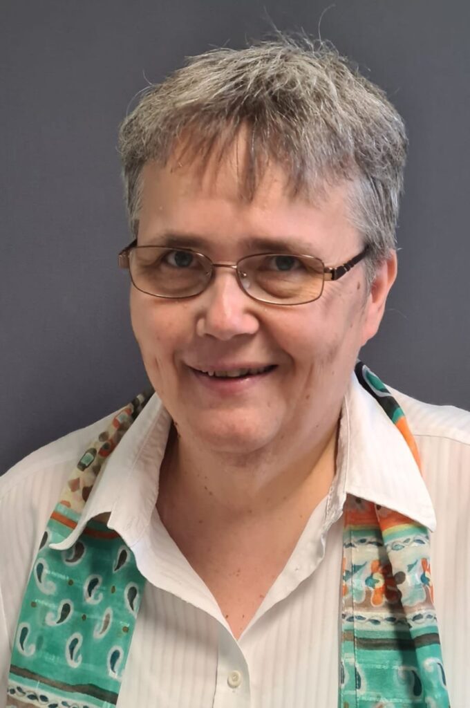 Eine Person mit Brille lächelt freundlich in die Kamera, trägt ein weißes Hemd und einen bunt gemusterten Schal.