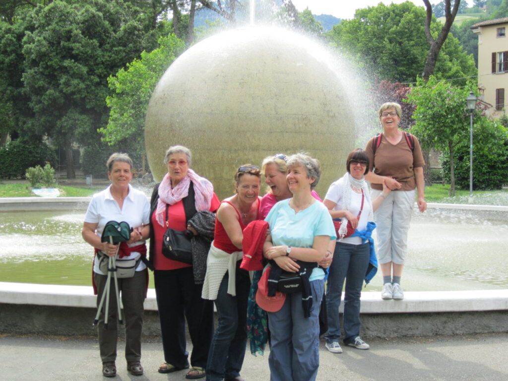 Eine Gruppe von lächelnden Frauen steht vor einem Brunnen mit einer großen Kugel in einem Park.