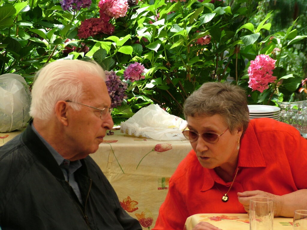 Ein älterer Mann und eine ältere Frau sitzen an einem Tisch im Freien und unterhalten sich, umgeben von blühenden Hortensien.