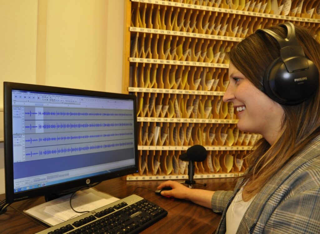 Eine Person mit Kopfhörern bearbeitet eine Audiodatei an einem Computer in einem Raum mit vielen kleinen Fächern im Hintergrund.