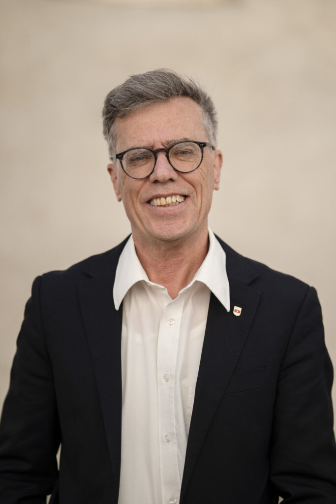 Ein lächelnder Mann mit Brille und grauem Haar trägt ein weißes Hemd und ein dunkles Sakko.