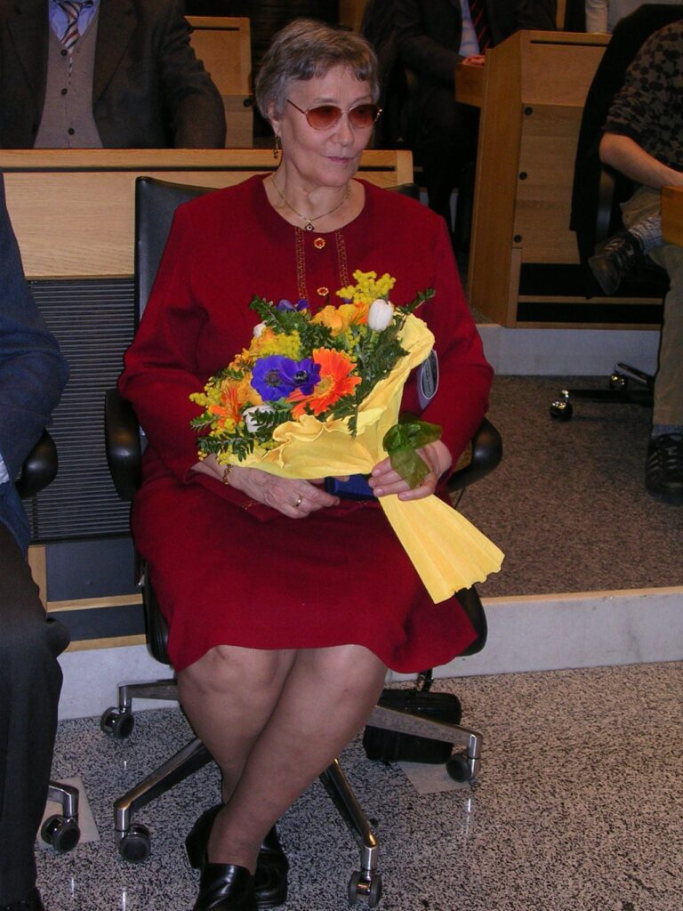 Eine Frau in rotem Kleid sitzt auf einem Stuhl und hält einen bunten Blumenstrauß in der Hand.