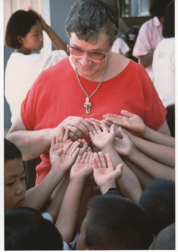 Eine ältere Person in roter Kleidung steht lächelnd inmitten einer Gruppe von Kindern, die ihre Hände ausstrecken.