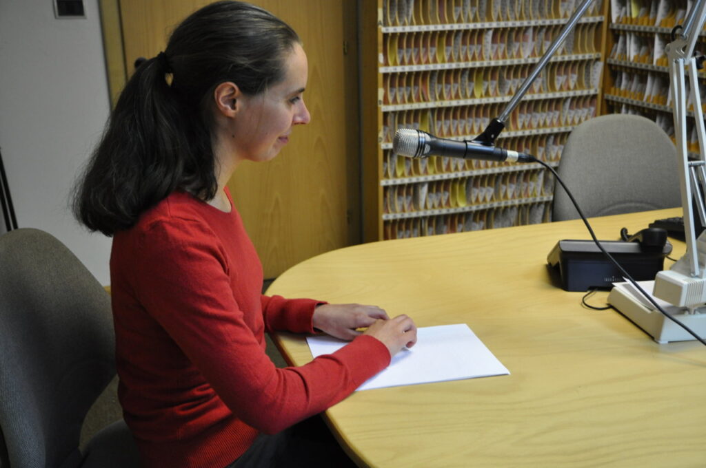 Eine Frau liest in Brailleschrift und spricht in ein Mikrofon an einem Tisch.