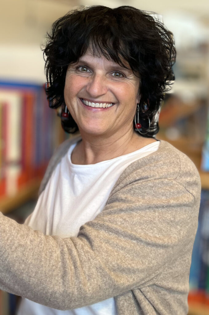 Eine lächelnde Person mit lockigem, dunklem Haar steht vor einem Bücherregal.