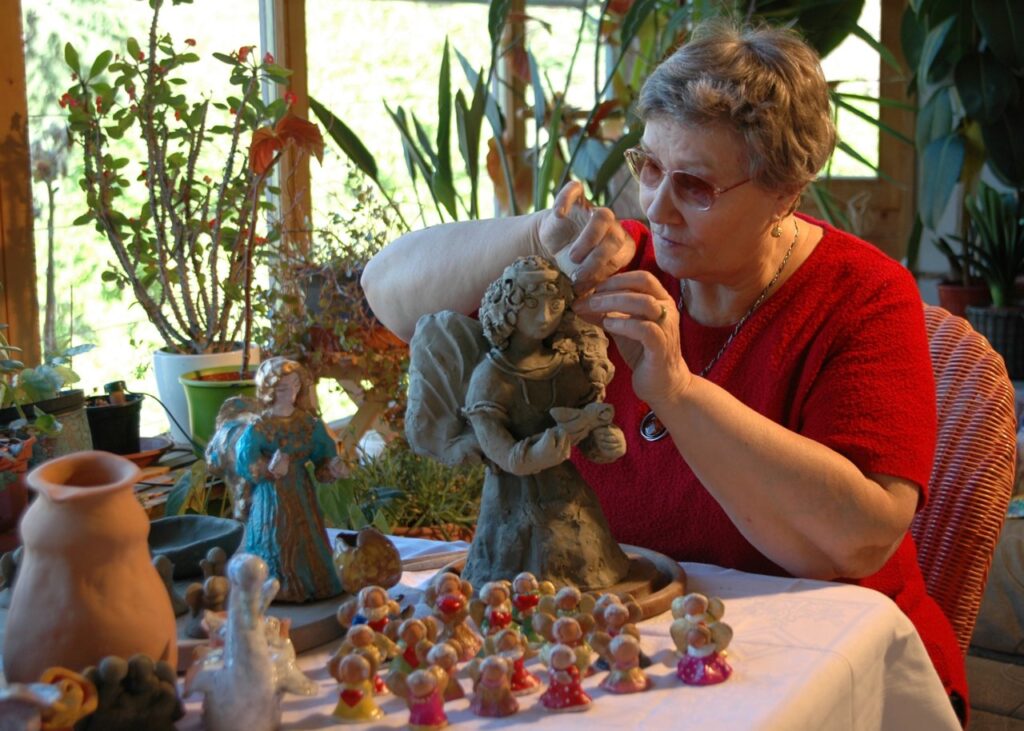 Eine Frau modelliert sorgfältig eine Engelsfigur aus Ton an einem Tisch mit weiteren kleinen Tonfiguren und Pflanzen im Hintergrund.