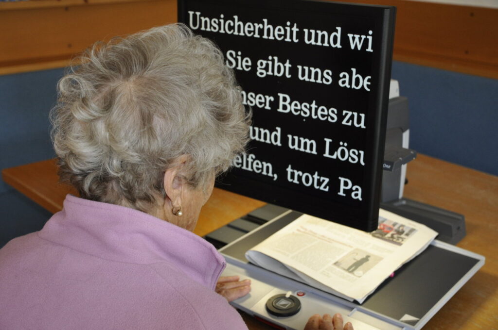 Eine ältere Frau liest mithilfe eines Vergrößerungsgeräts einen Text auf einem Bildschirm.