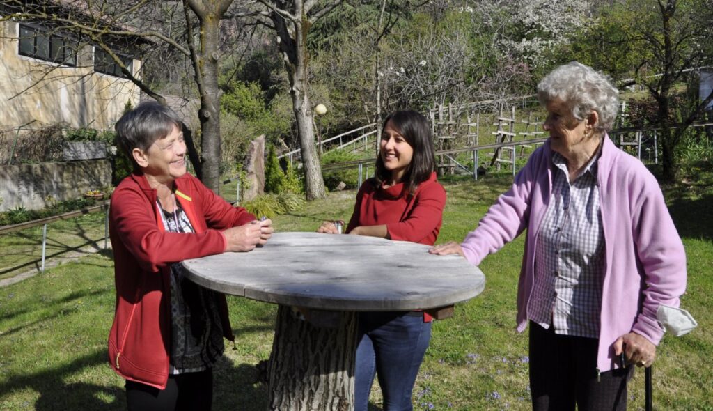 Drei Frauen stehen lächelnd um einen runden Holztisch im Garten.
