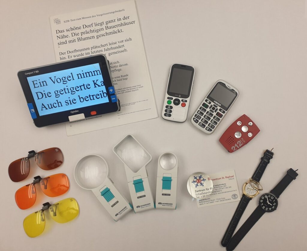 Hilfsmittel für sehbehinderte Menschen, darunter eine digitale Lupe, zwei einfache Mobiltelefone, farbige Bildschirmfilter, Handlupen, eine rote Sprachausgabe, eine Visitenkarte und zwei Uhren.