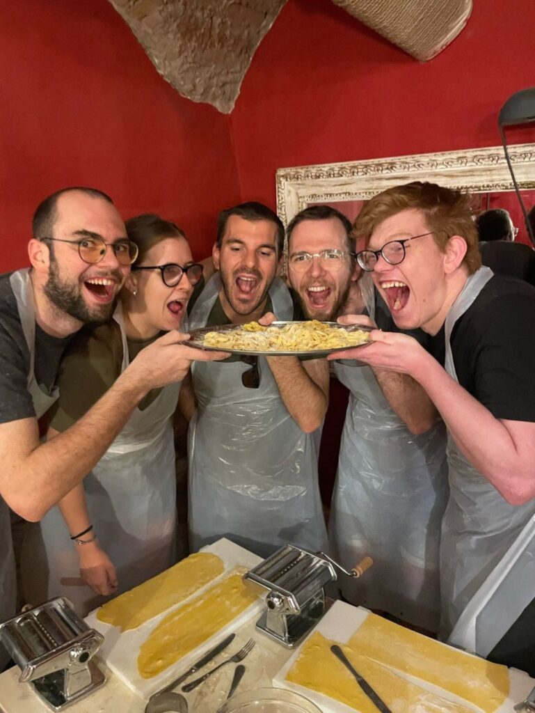 Eine Gruppe von fünf Personen in Schürzen hält freudig ein großes Teller mit frisch zubereiteten Nudeln und zeigt darauf.