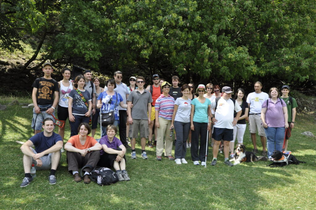 Eine Gruppe von Menschen steht und sitzt auf einer Wiese vor Bäumen, mit zwei Blindenhunden im Vordergrund.