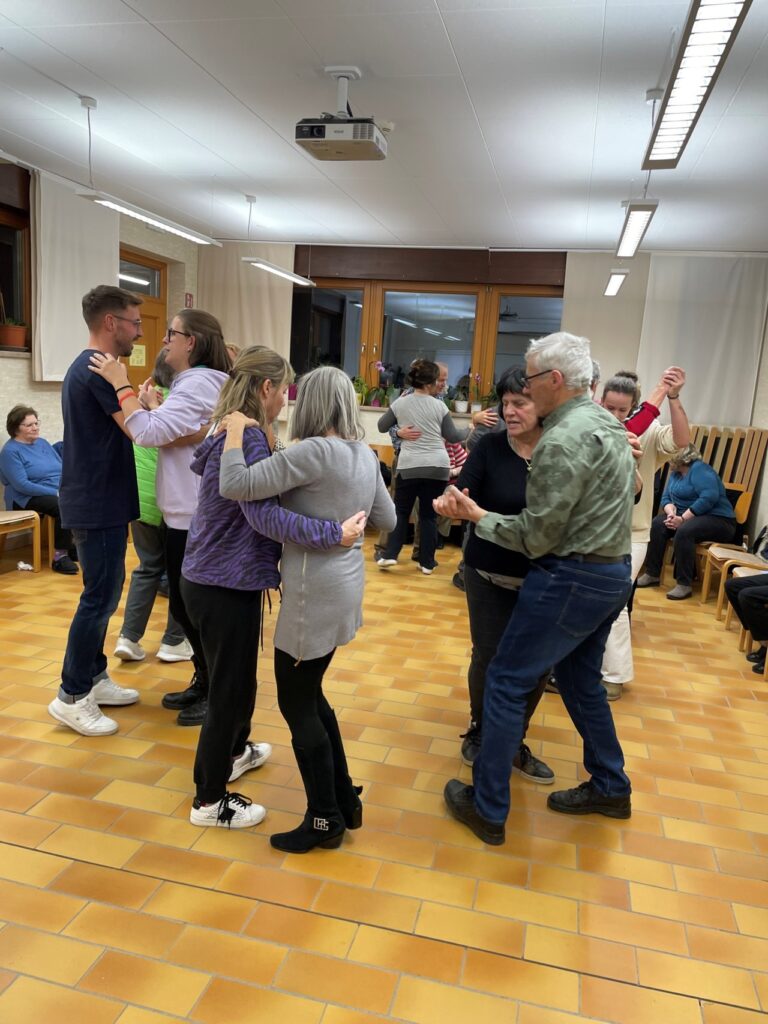 Mehrere Paare tanzen gemeinsam in einem hell erleuchteten Raum mit Holzboden.