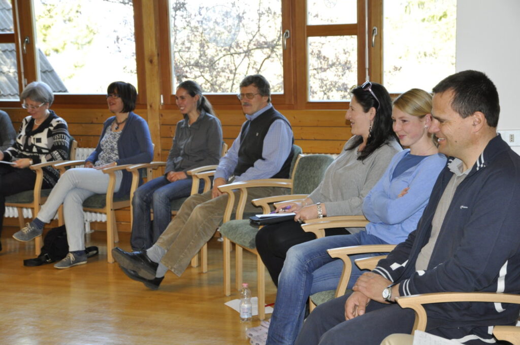 Eine Gruppe von lächelnden Menschen sitzt in einem Raum auf Stühlen in einer Reihe.