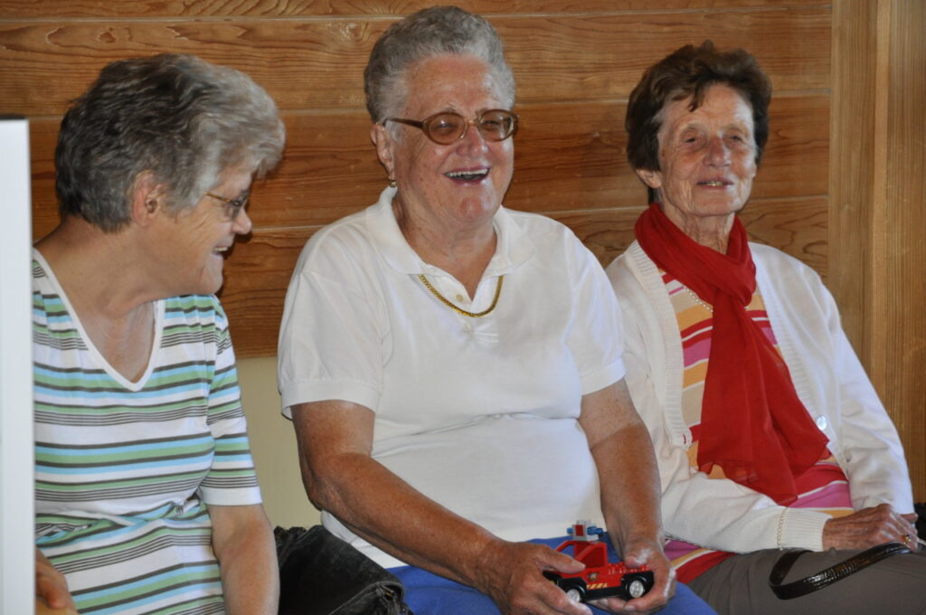 Drei ältere Frauen sitzen auf einer Bank und lachen, während eine von ihnen ein kleines rotes Spielzeugauto hält.
