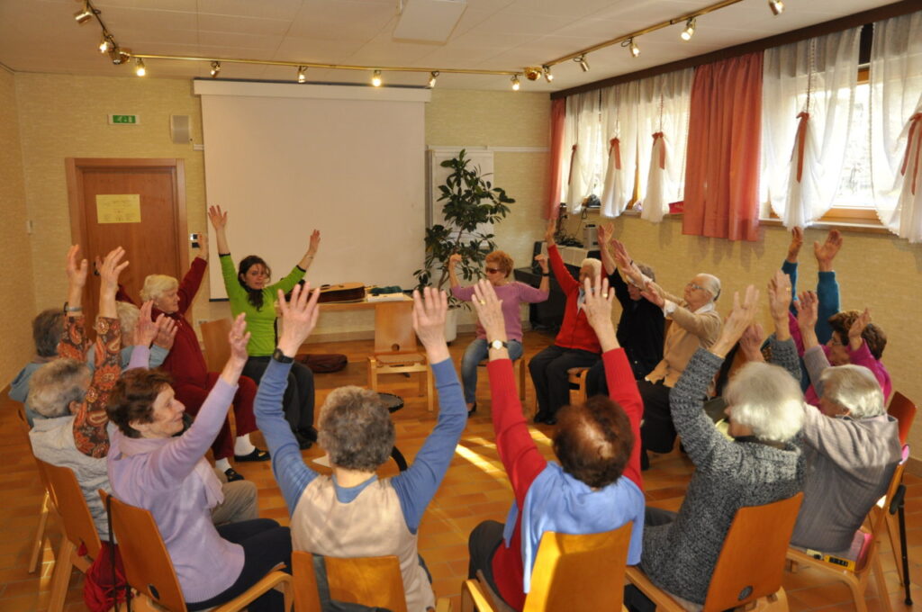 Eine Gruppe älterer Menschen sitzt im Kreis in einem Raum und hebt gemeinsam die Arme für eine Übung.