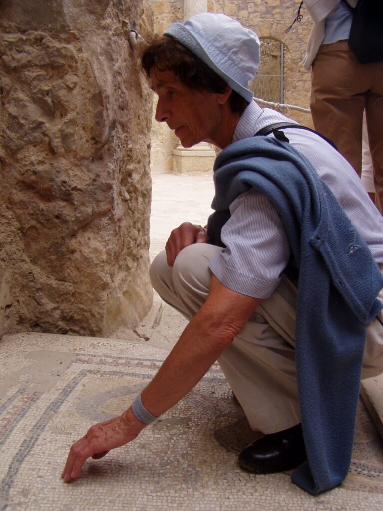 Eine Person im blauen Hut und Pullover berührt ein antikes Mosaik in einer archäologischen Stätte.