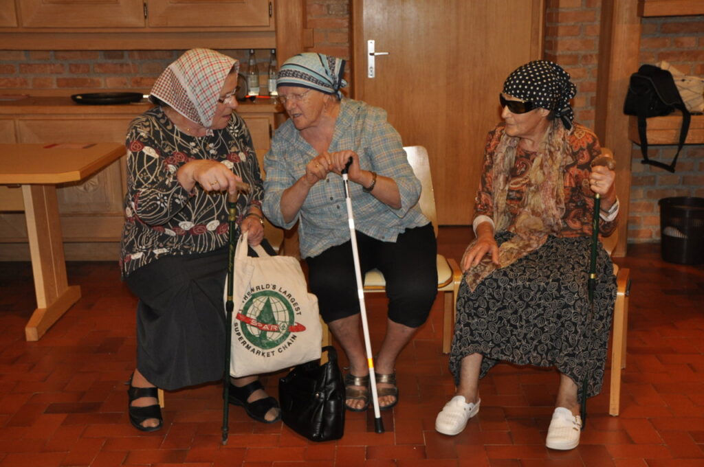 Drei ältere Frauen sitzen auf Stühlen, unterhalten sich und halten Spazierstöcke.