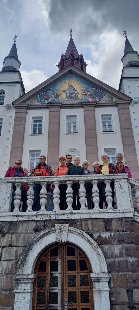 Eine Gruppe von Menschen steht auf einem Balkon vor einem historischen Gebäude mit kunstvoll gestaltetem Giebel, der religiöse Motive zeigt.