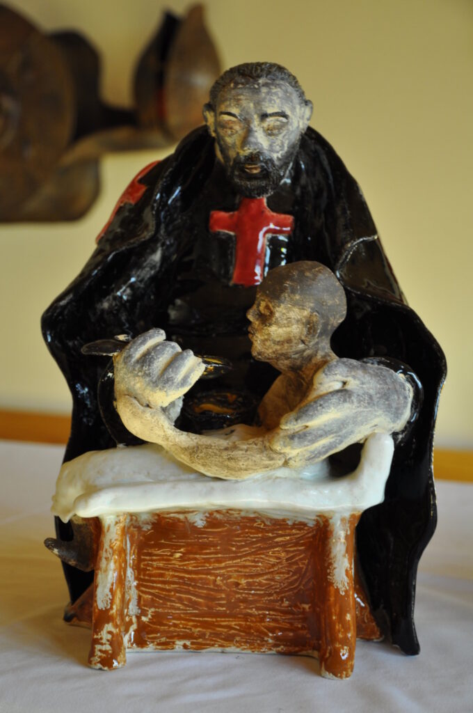 Eine Keramikskulptur zeigt eine sitzende Figur, die von einem in Schwarz gekleideten Mann mit rotem Kreuz umarmt wird.