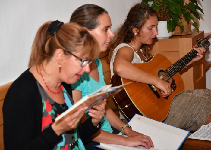 Drei Frauen sitzen zusammen, lesen aus Heften und eine spielt Gitarre.