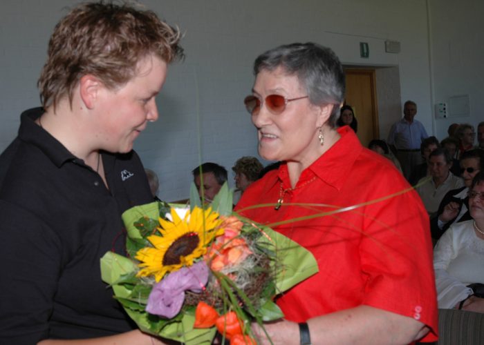 Eine Person in rotem Hemd lächelt und erhält einen Blumenstrauß von einer Person in schwarzem Poloshirt, während mehrere Menschen im Hintergrund zuschauen.