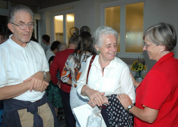 Eine Gruppe von älteren Personen unterhält sich fröhlich bei einer Veranstaltung in einem Raum mit Sonnblumen im Hintergrund.