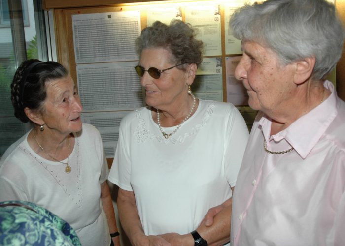 Drei ältere Frauen unterhalten sich lächelnd in einem Raum mit einer Pinnwand im Hintergrund.