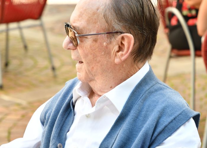 Ein älterer Mann im Profil sitzt draußen auf einem Stuhl, trägt eine Sonnenbrille und lächelt.