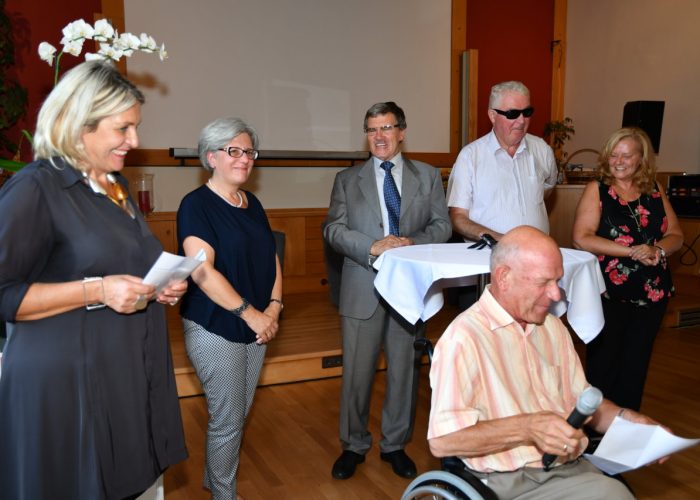 Eine Gruppe von Menschen steht lächelnd in einem Raum, eine Person sitzt im Rollstuhl und hält ein Mikrofon.