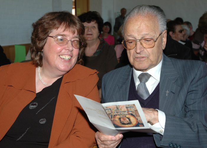 Eine lächelnde Frau und ein älterer Mann in einem Anzug betrachten gemeinsam ein Programmheft bei einer Veranstaltung.