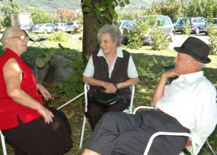 Drei ältere Menschen sitzen im Freien auf Stühlen und unterhalten sich im Schatten eines Baumes.