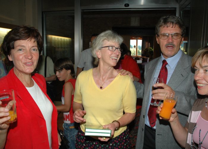 Eine Gruppe von Menschen unterschiedlichen Alters hält Getränke in der Hand und lächelt in einem Raum mit Glasfront.