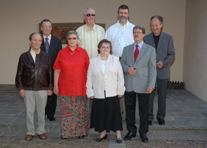 Eine Gruppe von acht älteren Menschen steht auf grauen Fliesen vor einem Gebäude und lächelt in die Kamera.