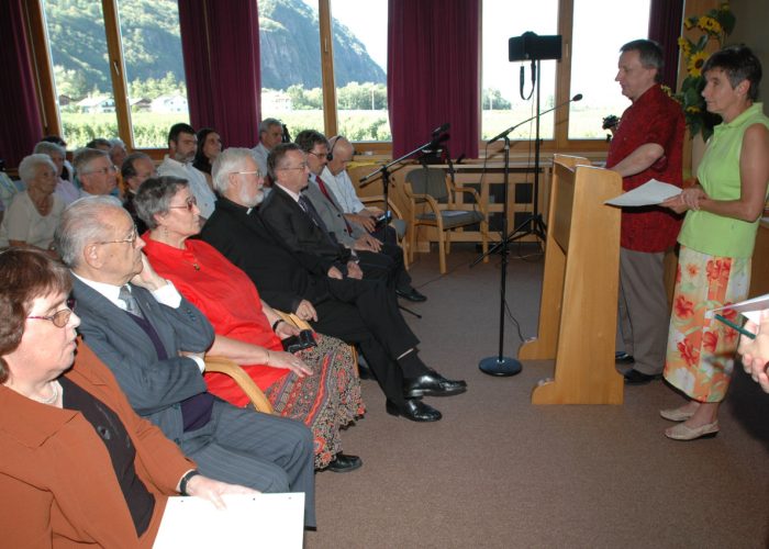 Eine Gruppe von Menschen sitzt auf Stühlen und schaut zu einem Rednerpaar am Pult in einem Raum mit großen Fenstern und violetten Vorhängen.
