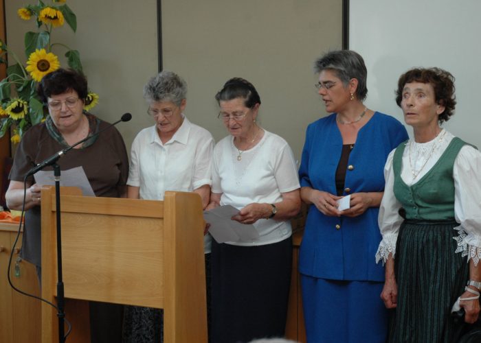 Fünf Frauen stehen nebeneinander und singen vor einem hölzernen Rednerpult mit Sonnenblumen im Hintergrund.
