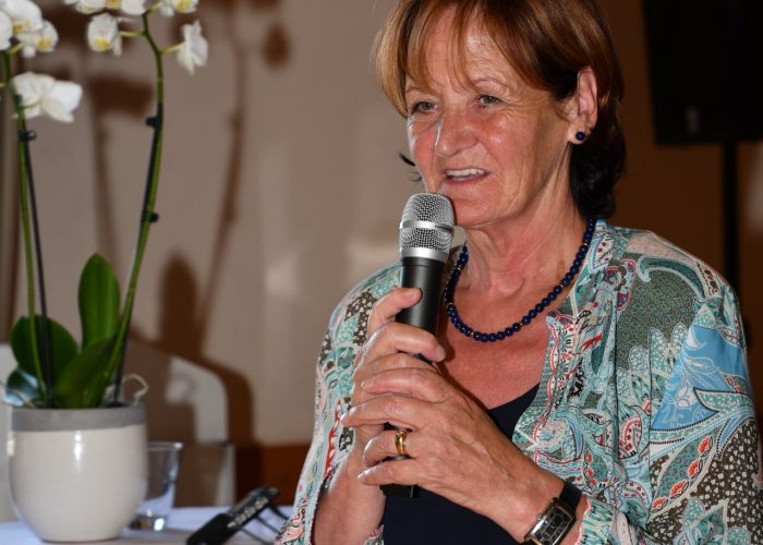 Eine Frau hält ein Mikrofon und spricht, neben ihr steht ein Tisch mit einer weißen Orchidee.