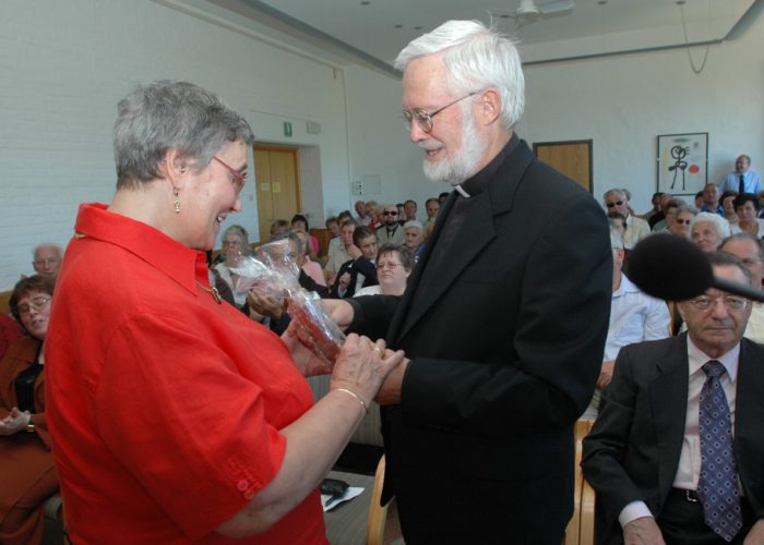 Eine Frau überreicht einem älteren Mann ein Geschenk, während viele Zuschauer im Hintergrund applaudieren.