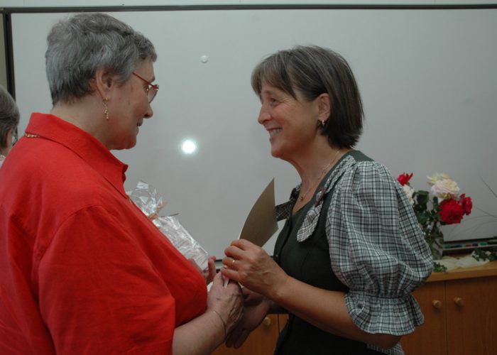 Zwei Frauen stehen sich lächelnd gegenüber und halten sich an den Händen, während sie miteinander sprechen, im Hintergrund sind Blumen.