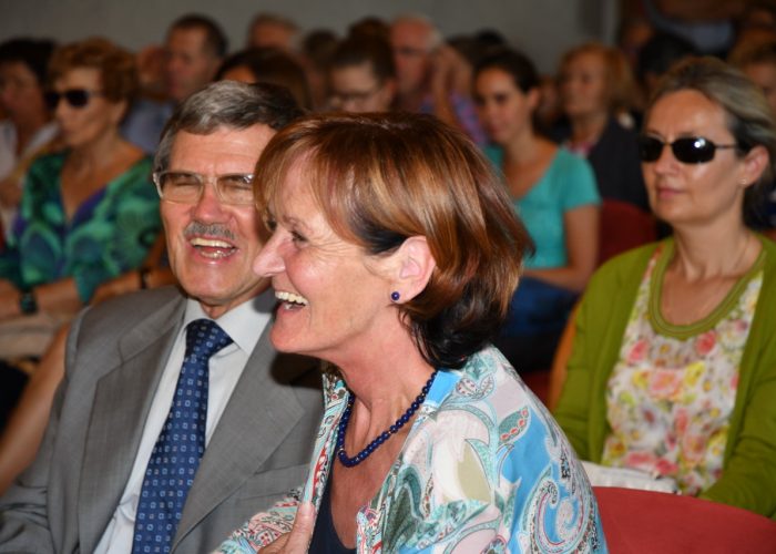 Ein lachendes älteres Paar sitzt in einem Publikum, umgeben von weiteren Personen.