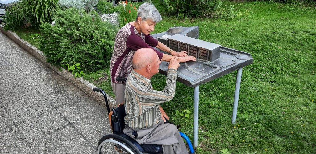 Eine blinde Frau und ein Rollstuhlfahrer erkunden ein Tastmodell im Freien